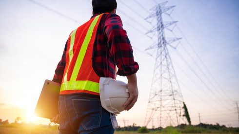 Engineer holding helmet and laptop in standard safety uniform working inspect the electricity high voltage pole